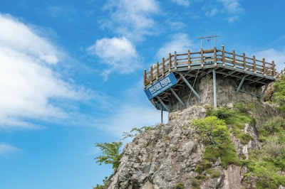 A wooden viewing platform is perched atop a rocky cliff, surrounded by lush green vegetation under a bright blue sky with scattered white clouds. A sign is attached to the platform.