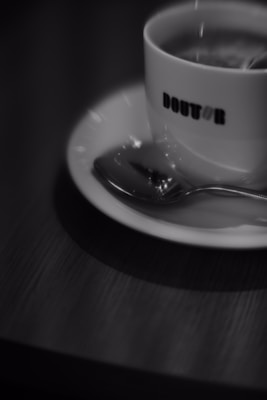 A cup of coffee sits on a saucer with a spoon next to it on a wooden table. The focus is slightly blurred, creating a soft and atmospheric feel. The word 'DOUTOR' is visible on the cup, which contains a steaming beverage.