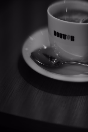 A cup of coffee sits on a saucer with a spoon next to it on a wooden table. The focus is slightly blurred, creating a soft and atmospheric feel. The word 'DOUTOR' is visible on the cup, which contains a steaming beverage.