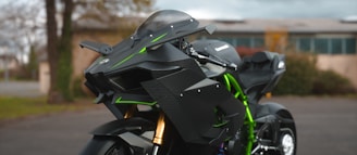 A sleek, modern motorcycle with an aggressive, angular design predominantly in black with green accents. The bike's front features advanced aerodynamics and prominent suspension components. Parked in an outdoor setting with blurred background, suggesting movement or speed.