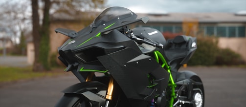 A sleek, modern motorcycle with an aggressive, angular design predominantly in black with green accents. The bike's front features advanced aerodynamics and prominent suspension components. Parked in an outdoor setting with blurred background, suggesting movement or speed.