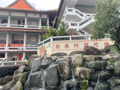 A multi-story building with traditional Asian architectural elements, featuring a red-tiled roof and ornate details. Stairs and greenery are prominent, and there's a decorative wall with Chinese characters and artwork. In the foreground, a small rock waterfall adds to the serene atmosphere.