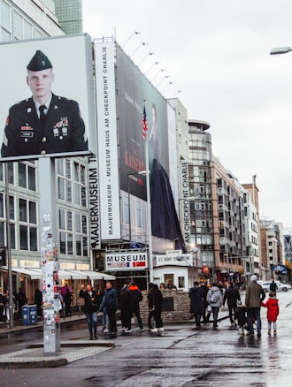 A city street scene with several pedestrians walking on wet pavement with buildings and large billboards in the background. One billboard features a person in military uniform and another displays an advertisement. Signs for the Mauer Museum and Checkpoint Charlie are visible, indicating a historical location.