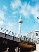 A tall, iconic television tower rises against a bright blue sky scattered with some clouds. Below, there's a railway bridge with overhead lines running across, and on the right, part of a modern glass structure is visible.