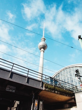 A tall, iconic television tower rises against a bright blue sky scattered with some clouds. Below, there's a railway bridge with overhead lines running across, and on the right, part of a modern glass structure is visible.