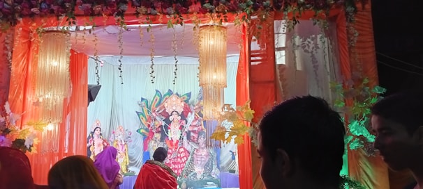 A decorated stage features vibrant and colorful statues of deities surrounded by flowers and ornate lights. People are gathered, possibly participating in a religious or cultural event.