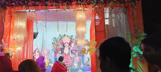 A vibrant orange-themed stage filled with devotees engaged in a bhakti event under soft glowing lights.
