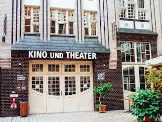 A building facade with art deco elements features a cinema and theater entrance. The architecture includes decorative brickwork and large windows. The signage reads 'KINO UND THEATER' above the entrance, and there is greenery in front with potted plants.