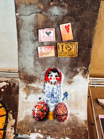 Street art featuring a Russian matryoshka doll painted on a textured, weathered wall. The doll has a white body with intricate blue designs resembling architectural elements and a red head with a cheerful face. Surrounding the doll are colorful cut-out letters spelling humorous or edgy phrases. The wall displays signs with bold messages in a mix of pink, red, and yellow.