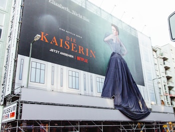 A large outdoor billboard advertising a Netflix series titled 'Die Kaiserin.' The billboard features a striking image of a woman in a dramatic, flowing dress. The backdrop of the billboard mimics the façade of a building with window-like designs. A lamppost is visible in the foreground, slightly obstructing part of the view.