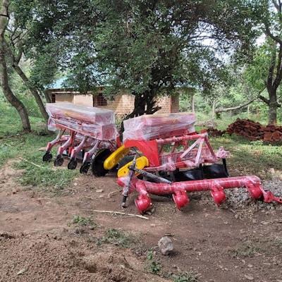 Large, colorful agricultural machinery covered in plastic sits on a dirt patch surrounded by greenery and trees. A small brick building is partially visible in the background.