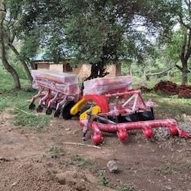 Large, colorful agricultural machinery covered in plastic sits on a dirt patch surrounded by greenery and trees. A small brick building is partially visible in the background.