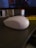 A white computer mouse is placed on a dark, textured wooden desk surface. The backdrop includes an out-of-focus monitor playing a video and some office supplies like a pen holder and a yellow book.