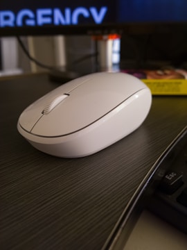 A white wireless computer mouse is placed on a dark wooden desk. The background includes a blurred computer screen displaying the partial word 'EMERGENCY' along with some other indistinct objects.