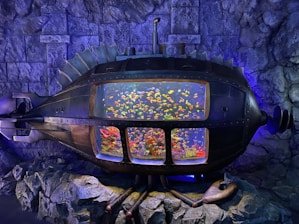 An intricately designed metallic submarine-like structure embedded in rock walls, featuring a large, transparent display in the center showcasing a vibrant aquarium filled with colorful fish and corals. The surrounding area is dimly lit with a bluish hue, highlighting the details of the structure.