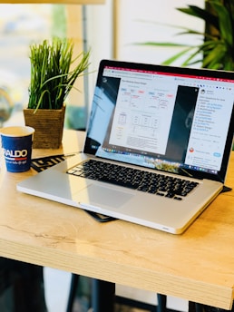 A laptop displaying a webpage about non-blocking queue design is placed on a wooden table. Next to it is a potted plant and a disposable coffee cup with branding. The setup is near a window, suggesting a cozy or casual workspace environment.