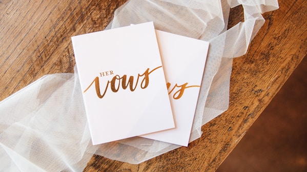 white cards saying his and her vows in gold script lettering on wooden table with white lace 