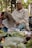 An elderly man sits outdoors, reading a newspaper. He is wearing a light-colored long-sleeve shirt and checkered pants. In front of him, there is a variety of fresh vegetables, including bananas and leafy greens. Behind him, a street scene with trees and vehicles is partially visible.
