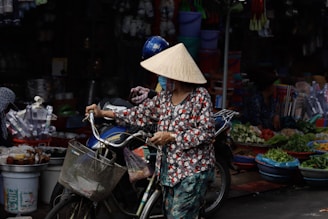 Local woman wearing a Non La conical hat pushing a bicycle with a basket in a Vietnamese market