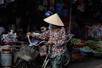 A person wearing a traditional conical hat is pushing a bicycle through a market. The market is vibrant, with various vegetables and goods visible in the background. The person is dressed in colorful clothing and appears to be carrying something in the bicycle's front basket.