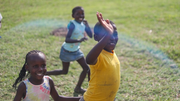 Three children are playing joyfully on a grassy field, with one child in the foreground wearing a yellow shirt and smiling while waving at the camera. The other two children are in motion in the background, also smiling and enjoying the moment.