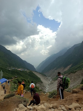 Hikers are seen descending a mountain trail with a dramatic landscape of lush green valleys and towering mountains. Above, a vast sky is filled with dynamic, swirling clouds revealing patches of blue. The pathway is busy with people carrying backpacks and walking sticks, suggesting a popular trekking route.