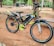 A brightly colored black and green bicycle is parked on a sandy surface. The bicycle features distinct neon yellow accents on the pedals, rims, and frame. It is equipped with sturdy tires suitable for off-road use. The background includes a park-like setting with trees, a bench, and a few parked vehicles.