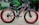 A black electric bicycle with thick red-rimmed tires is parked on a paved path. The bicycle bears the brand name 'VOLTEBYK' in red on its frame. Behind it, green foliage and signs with text are visible.