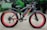 A black electric bicycle with thick red-rimmed tires is parked on a paved path. The bicycle bears the brand name 'VOLTEBYK' in red on its frame. Behind it, green foliage and signs with text are visible.