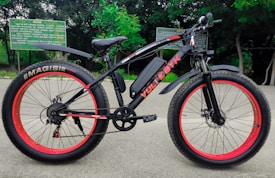 A black electric bicycle with thick red-rimmed tires is parked on a paved path. The bicycle bears the brand name 'VOLTEBYK' in red on its frame. Behind it, green foliage and signs with text are visible.