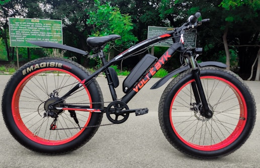 A black electric bicycle with thick red-rimmed tires is parked on a paved path. The bicycle bears the brand name 'VOLTEBYK' in red on its frame. Behind it, green foliage and signs with text are visible.