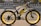 A black and yellow electric bicycle with thick tires is parked on the side of a paved road. The bicycle features branding and text on the frame and wheels, and has a sturdy, modern design. It is located in an urban setting with a road and buildings in the background.