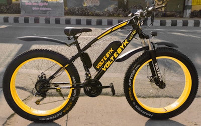 A black and yellow electric bicycle with thick tires is parked on the side of a paved road. The bicycle features branding and text on the frame and wheels, and has a sturdy, modern design. It is located in an urban setting with a road and buildings in the background.