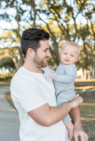 A man with dark hair and a beard wears a white shirt and holds a baby dressed in a gray outfit. They are outdoors, surrounded by a park-like setting with trees and soft sunlight filtering through the branches. Both appear to be smiling and enjoying a moment together.