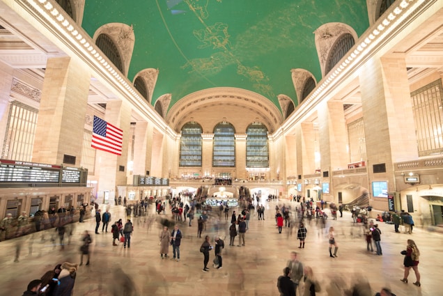 A large, bustling train station with high arched ceilings featuring an intricate celestial mural. The spacious hall is illuminated with soft lighting from elegant fixtures and a prominently displayed American flag. Numerous travelers and commuters can be seen moving through the space, creating a sense of constant motion and activity.