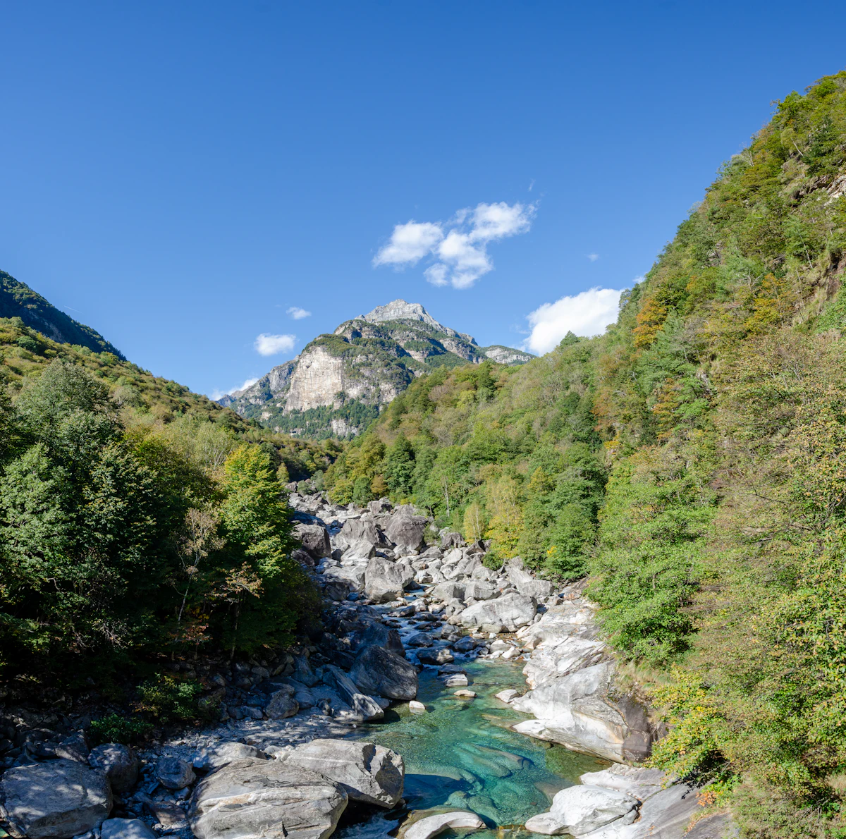 The Verzasca river running over pale boulders between wooded slopes, below a rocky peak