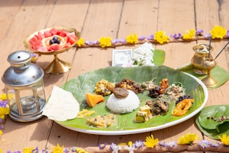 A traditional meal is presented on a large banana leaf placed on a plate, with an assortment of various vegetables, curries, and rice. A piece of flatbread is also on the leaf. To the left, there is a metallic lantern. In the background, there's a golden bowl containing cut fruit like watermelon and berries, and a brass container is on the right side along with incense sticks emitting smoke. The entire arrangement is bordered with a garland of yellow and purple flowers, and a single dollar bill is placed near the top.