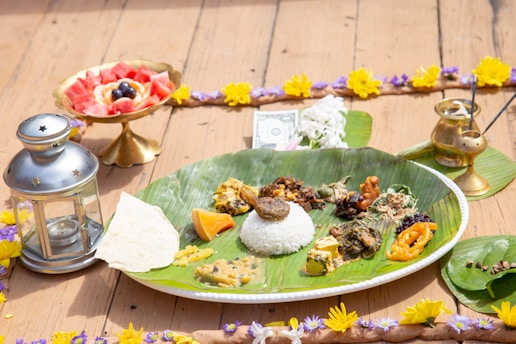 A traditional meal is presented on a large banana leaf placed on a plate, with an assortment of various vegetables, curries, and rice. A piece of flatbread is also on the leaf. To the left, there is a metallic lantern. In the background, there's a golden bowl containing cut fruit like watermelon and berries, and a brass container is on the right side along with incense sticks emitting smoke. The entire arrangement is bordered with a garland of yellow and purple flowers, and a single dollar bill is placed near the top.
