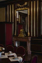 An opulent interior featuring a wooden wall adorned with gold accents and a large ornate mirror. A lavish mantle holds a decorative clock and sculpture. In the foreground, a table set with teacups and saucers on lace placemats is surrounded by upholstered chairs.