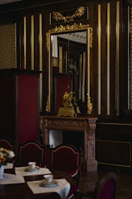 An opulent interior featuring a wooden wall adorned with gold accents and a large ornate mirror. A lavish mantle holds a decorative clock and sculpture. In the foreground, a table set with teacups and saucers on lace placemats is surrounded by upholstered chairs.
