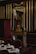 An opulent interior featuring a wooden wall adorned with gold accents and a large ornate mirror. A lavish mantle holds a decorative clock and sculpture. In the foreground, a table set with teacups and saucers on lace placemats is surrounded by upholstered chairs.