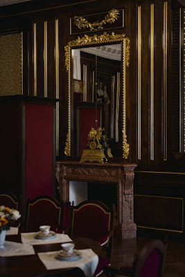 An opulent interior featuring a wooden wall adorned with gold accents and a large ornate mirror. A lavish mantle holds a decorative clock and sculpture. In the foreground, a table set with teacups and saucers on lace placemats is surrounded by upholstered chairs.