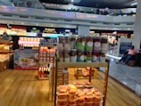 A food stall in an airport terminal displaying a variety of packaged snacks including jars of cookie sticks, boxes of sweets, and bottles of condiments. The stall is well-lit and situated near other stores. People are visible on the upper level balcony, and a seated person is on the right side.