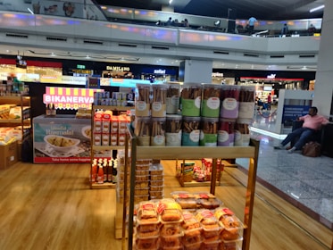 A food stall in an airport terminal displaying a variety of packaged snacks including jars of cookie sticks, boxes of sweets, and bottles of condiments. The stall is well-lit and situated near other stores. People are visible on the upper level balcony, and a seated person is on the right side.