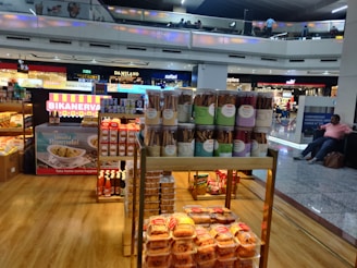 A food stall in an airport terminal displaying a variety of packaged snacks including jars of cookie sticks, boxes of sweets, and bottles of condiments. The stall is well-lit and situated near other stores. People are visible on the upper level balcony, and a seated person is on the right side.
