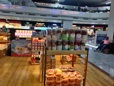 A food stall in an airport terminal displaying a variety of packaged snacks including jars of cookie sticks, boxes of sweets, and bottles of condiments. The stall is well-lit and situated near other stores. People are visible on the upper level balcony, and a seated person is on the right side.