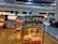 A food stall in an airport terminal displaying a variety of packaged snacks including jars of cookie sticks, boxes of sweets, and bottles of condiments. The stall is well-lit and situated near other stores. People are visible on the upper level balcony, and a seated person is on the right side.