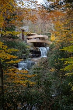 A modern architectural structure, blending harmoniously with its natural surroundings, is perched over a flowing waterfall. The scene is enveloped by dense woodland showcasing vibrant autumn foliage. The building features horizontal lines and balconies with people visible on one of the terraces, highlighting its integration with the environment.