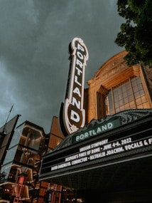 A vintage theater marquee with 'Portland' prominently displayed in illuminated letters, set against a cloudy sky. The building features ornate architectural details with a mix of modern glass elements. Trees frame the right side, adding greenery to the urban scene. A sign lists details of a symphony event.