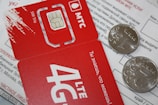A close-up of a red SIM card holder from MTS with its accompanying packaging. The card displays '4G LTE' and includes a microchip design. Two coins are placed on top of a printed document, partially obscuring the text in Russian.